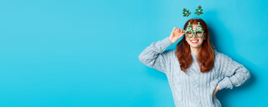 Winter holidays and Christmas sales concept. Beautiful redhead female model celebrating New Year, wearing funny party headband and glasses, smiling at camera.