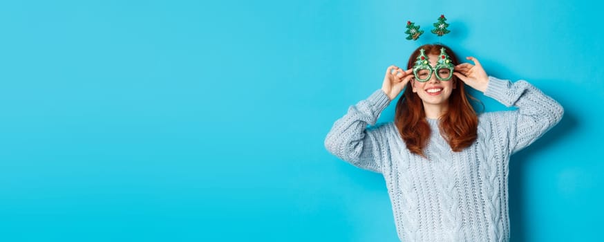 Winter holidays and Christmas sales concept. Beautiful redhead female model celebrating New Year, wearing funny party headband and glasses, smiling silly, blue background.