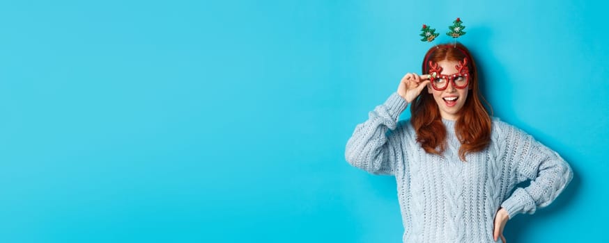 Christmas party and celebration concept. Cute redhead teen girl celebrating New Year, wearing xmas tree headband and funny glasses, looking left amused, blue background.