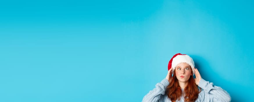 Winter holidays and Christmas eve concept. Head of funny redhead girl in santa hat, appear from bottom and squinting, making silly faces, standing near copy space on blue background.
