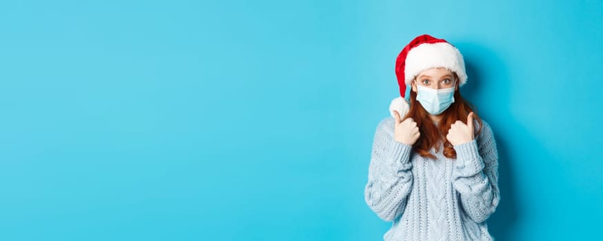 Christmas, quarantine and covid-19 concept. Cute teen redhead girl in santa hat and sweater, wearing face mask from coronavirus, showing thumbs up, standing over blue background.