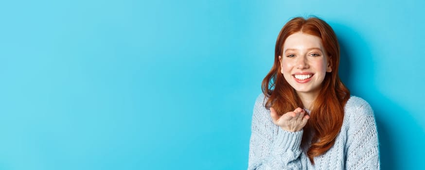 Lovely redhead female model smiling, sending air kiss at camera, standing against blue background.