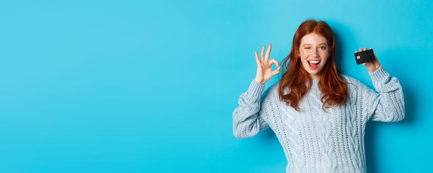 Happy redhead girl in sweater showing credit card and okay sign, recommending bank offer, standing over blue background.