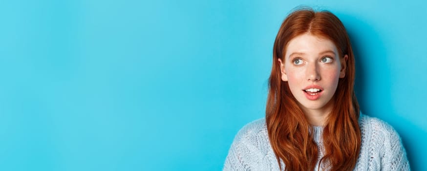 Headshot of thoughtful redhead teen girl looking at upper left corner, staring at logo with curious expression, standing over blue background.