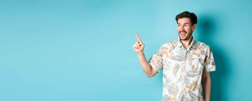 Excited smiling tourist pointing and looking aside at empty space, checking out promo offer, standing in hawaiian shirt on blue background.