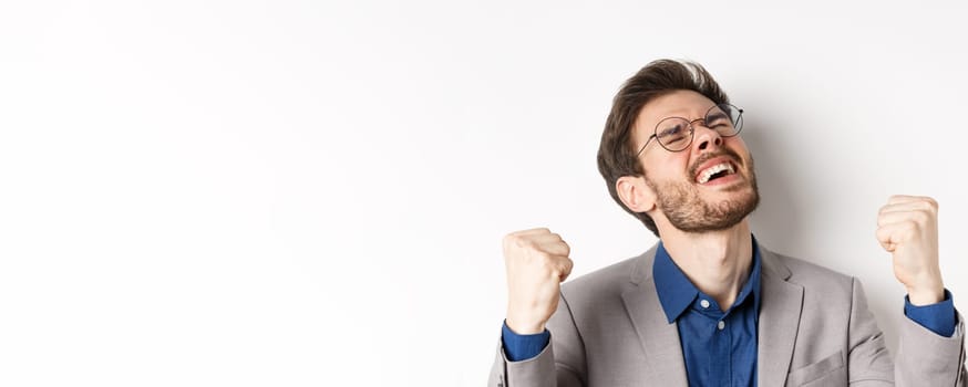 Excited businessman in glasses and suit shouting yes with pleasure and relieved face, shaking fists up, triumphing, winning bet, standing on white background.