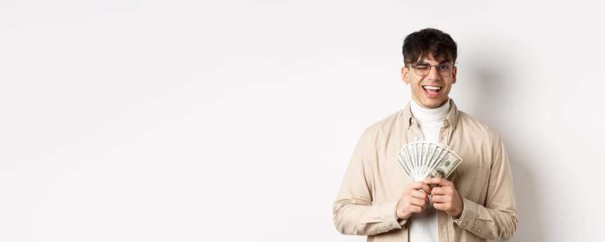 Handsome young man winking and showing dollar bills, holding fan of money banknotes and smiling pleased, making money, standing on white background.