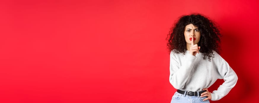 Angry young woman with curly hair and red lips, frowning and shushing, tell to be quiet, make taboo gesture to keep mouth shut, standing on red background.