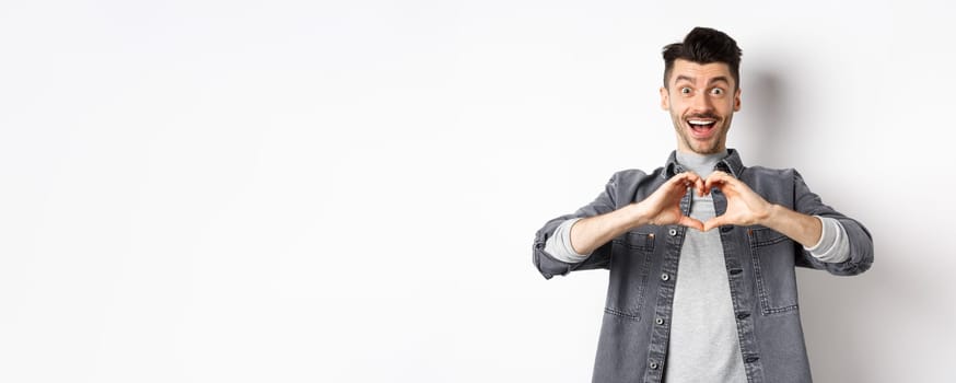 Romantic boyfriend showing heart gesture and looking with love at camera, celebrating White day after valentines, standing on white background. Romance concept.