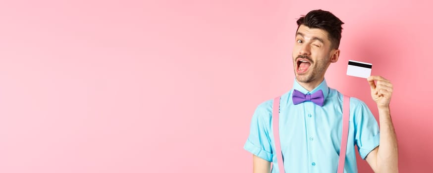 Shopping concept. Funny guy with moustache winking at camera, showing plastic credit card, recommending bank promo offer, standing on pink background.