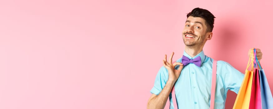 Confident smiling guy adjusting his bow-tie and showing gifts in shopping bags, looking happy, standing over pink background.