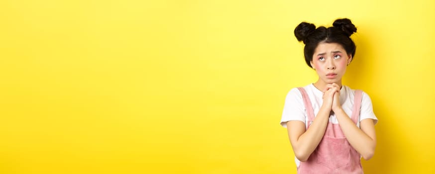 Sas asian girl holding hands in pray, pleading god, looking up and begging, standing on yellow background.