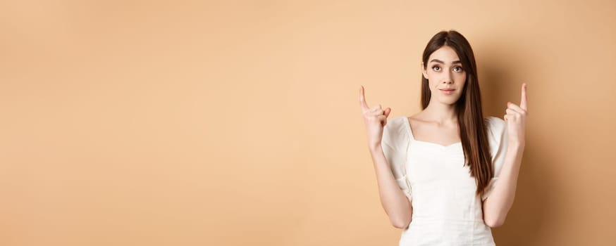 Hopeful romantic girl in white dress pointing fingers up, showing empty space for company logo, standing on beige background.