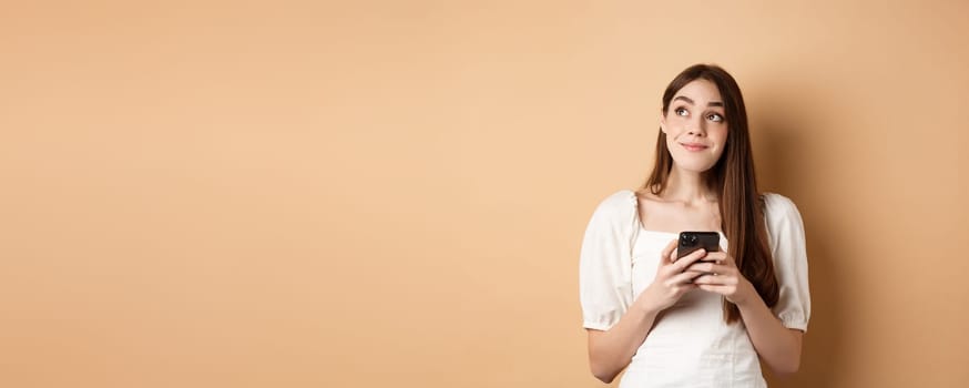 Romantic girl thinking with phone, looking aside and smiling dreamy, using dating app on smartphone, standing on beige background.