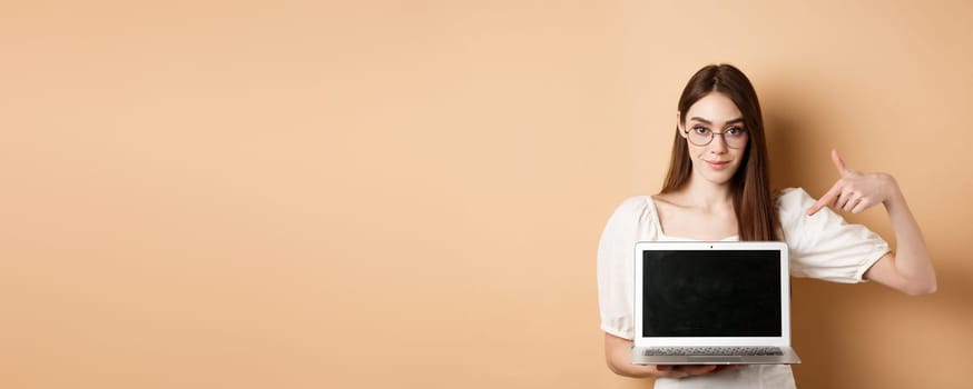 Confident young woman in glasses pointing at laptop screen, showing computer display, standing on beige background.