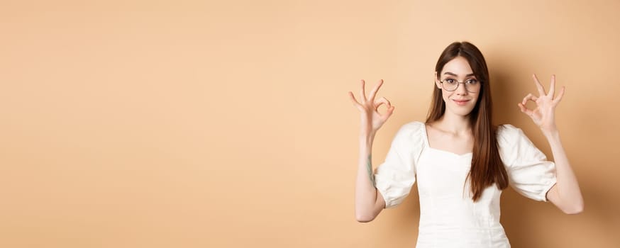 Satisfied female model in glasses show okay sign, looking pleased, agree and approve, standing on beige background.