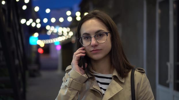 A young woman talks on the phone late at night on a narrow street. A girl with glasses communicates on a mobile phone. 4k