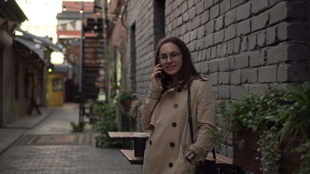 A young woman talks on the phone while standing against a wall on a narrow street. A girl with glasses and a coat speaks on the phone. 4k