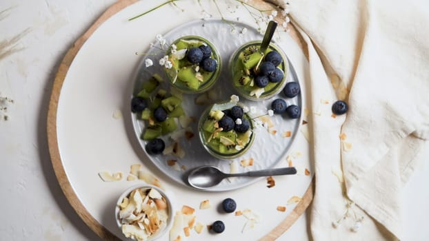 Chia pudding with kiwi, blueberries and coconut slices, three portions in glass jars on a white table.