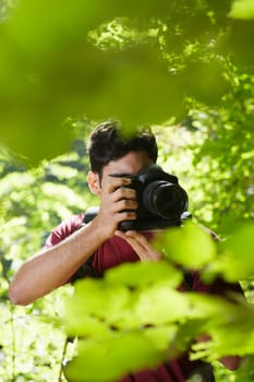 young hispanic man trekking among trees and taking pictures with dslr camera. Vertical shape, front view, copy space