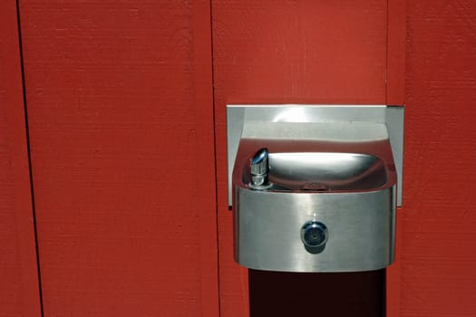 Silver steel water fountain attached to a painted red wood wall outdoors on a sunny day.