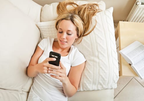 Pretty young blond woman lying on sofa and messaging with her smartphone