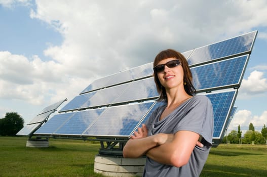 Wide angle torso shot of a young brunette woman in casual casual dress with crossed arms in front of photovoltaic system on green meadow and cloudy sky in summer