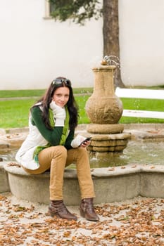 Autumn park young woman dialing phone sitting on fountain