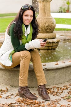 Autumn park young woman dialing phone sitting on fountain
