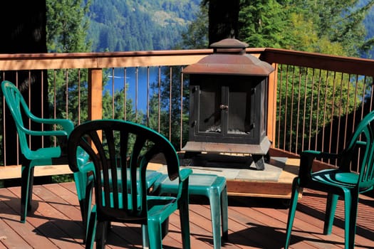 Four plastic chairs on a scenic sunny outdoor deck with a fireplace on a raised platform and a small table all surrounded by a wood and metal railing.