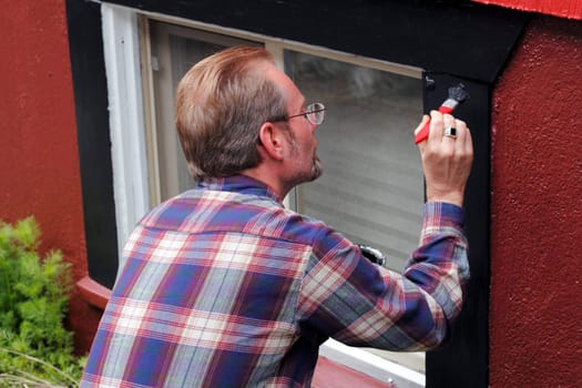 Man painting house window trim with black paint outdoors in the day.