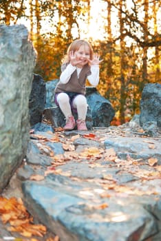 Small girl (3 years old) with smile is sitting on big rock and showing open hands. She plays in the autumn park at the evening