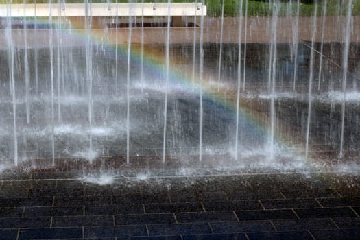 Water jets spraying H20 up out of paver bricks on a bright sunny day causing a rainbow to appear in the water mist in a public park.