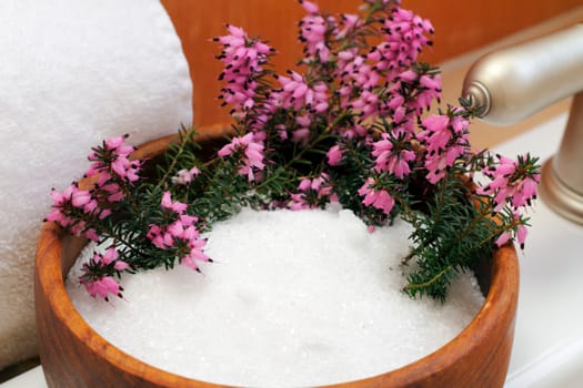 Pink heather flowers in a wood bowl filled with Epsom salts on the top side of a white bath tub near a white cotton towel and silver faucet.