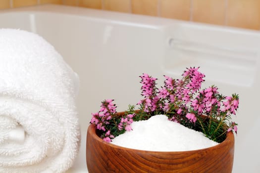 Fresh pink purple heather flowers in a teak wood bowl with epsom salts on the edge of a bathtub with a rolled up white towel ready to take a relaxing bath.