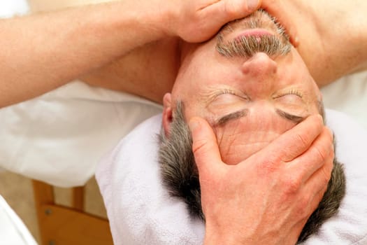 Mature Caucasian man in his 50s relaxing on a massage table with his eyes closed receiving a face massage from a massage therapist.