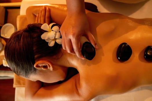 Closeup hot stone massage at spa salon in luxury resort with warm candle light, blissful woman customer enjoying spa basalt stone massage glide over body with soothing warmth. Quiescent