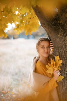 The blonde stands near the autumn tree. Thoughtful woman looks ahead, dressed in a yellow dress. Autumn content.