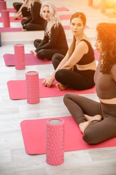 A group of six athletic women doing pilates or yoga on pink mats in front of a window in a beige loft studio interior. Teamwork, good mood and healthy lifestyle concept