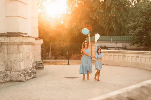 Daughter mother run holding hands. In blue dresses with flowing long hair, they hold balloons in their hands against the backdrop of a sunset and a white building