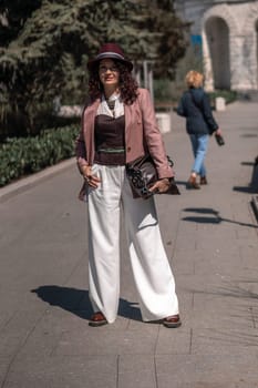 Woman park city. Stylish woman in a hat walks in a park in the city. Dressed in white corset trousers and a pink jacket with a bag in her hands