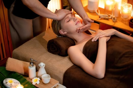 Caucasian woman enjoying relaxing anti-stress head massage and pampering facial beauty skin recreation leisure in warm candle lighting ambient salon spa in luxury resort or hotel. Quiescent