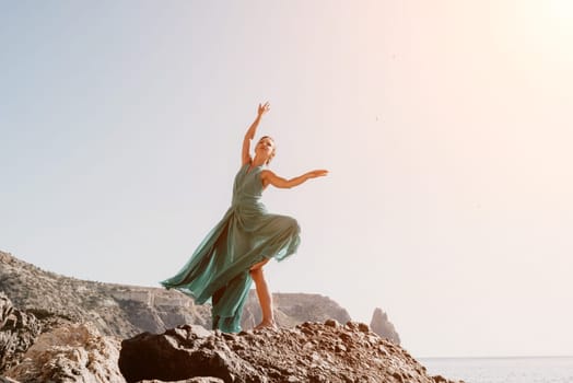 Side view a Young beautiful sensual woman in a red long dress posing on a rock high above the sea during sunrise. Girl on the nature on blue sky background. Fashion photo.