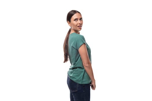 30 year old slender European brunette woman with a ponytail hairstyle dressed in a green T-shirt stands sideways on a white background with copy space.