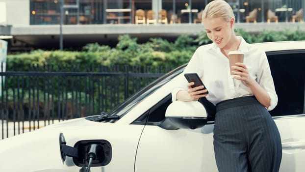 Progressive businesswoman leaning on electric car and charging station before driving around city center. Eco friendly rechargeable EV car powered by sustainable and clean energy.