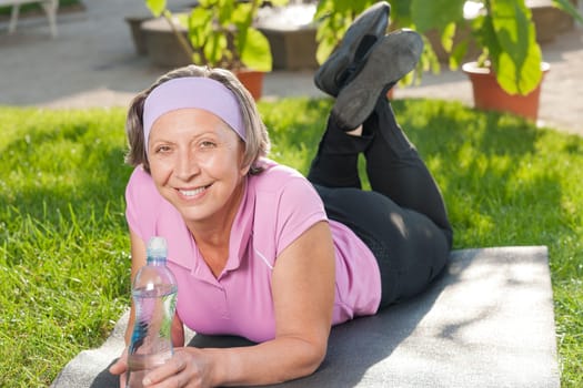 Senior sportive woman lying on mat drink water outside sunny day