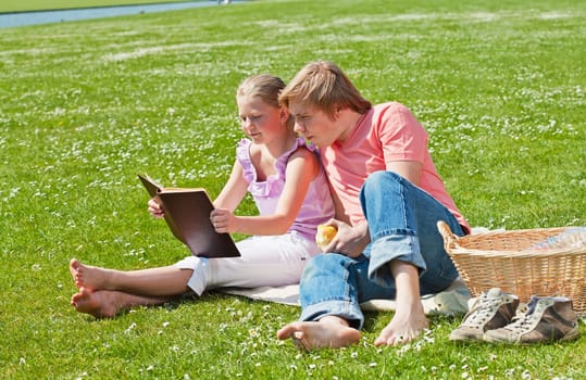 Teenager siblings reading book at picnic in the park in a bright sunny day