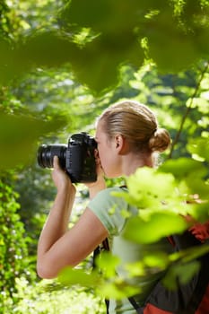 young woman trekking among trees and taking pictures with dslr camera. Vertical shape, side view, waist up, copy space