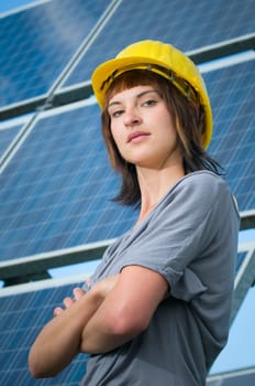 Side torso shot of a young brunette woman with a yellow construction helmet,casual casual clothes and crossed arms smiling contentedly into the camera in front of a free-standing photovoltaic system