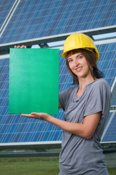 Upper body shot of a young brunette woman in casual casual dress,yellow construction helmet and green area in her hands in front of free-standing photovoltaic system